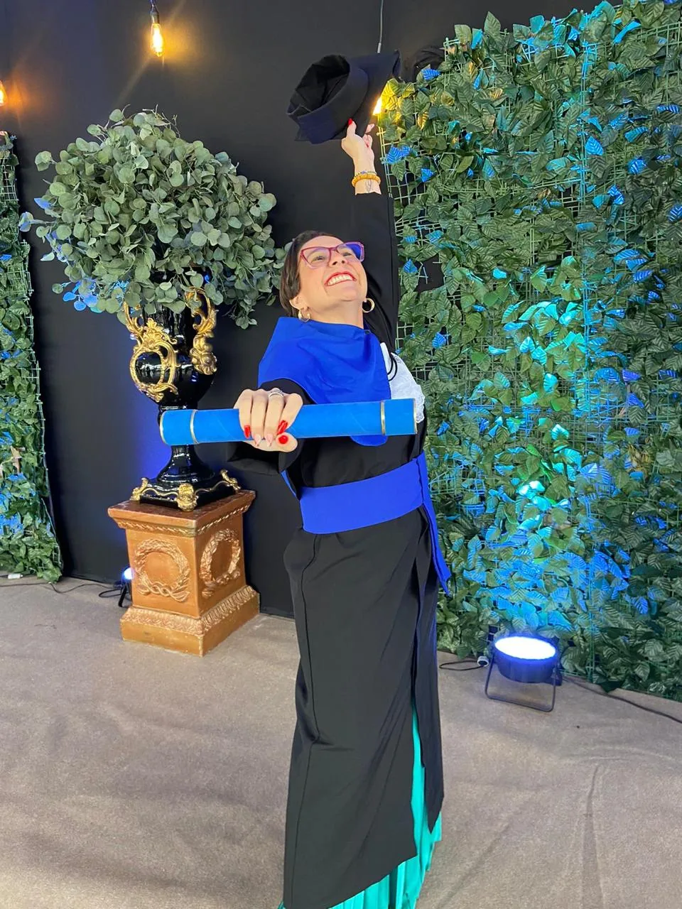Formanda de beca preta com faixa azul segurando o canudo e erguendo o capelo